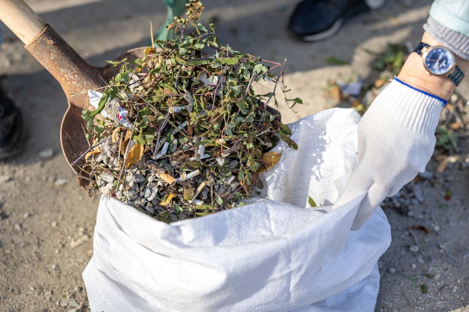 A large pile of mixed household waste and rubbish, including cardboard boxes, plastic bags, paper debris, and discarded packaging, cluttering a city pavement next to two overflowing wheelie bins, one